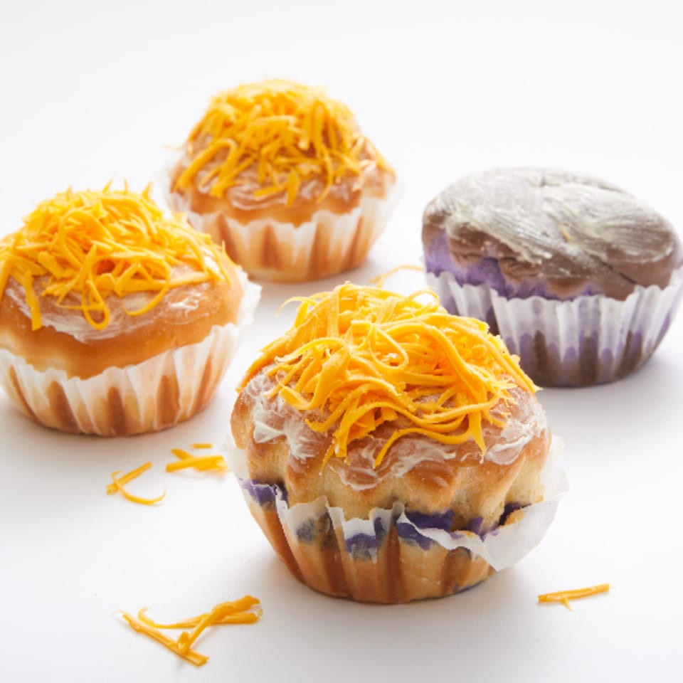 Four Goldilocks mamon, three topped with cheese and one with ube, on a white surface.