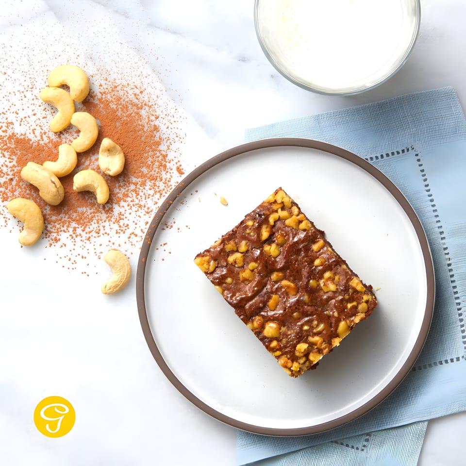 A Goldilocks cashew brownie on a white plate, with cashews, cocoa powder, and milk.
